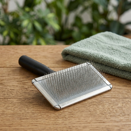 Brosse carde pour chien à picots fins en acier inoxydable et manche noir, posée près d'une serviette